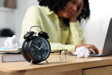 Deadline concept. Black alarm clock near woman working at wooden desk indoors, selective focus. Space for text