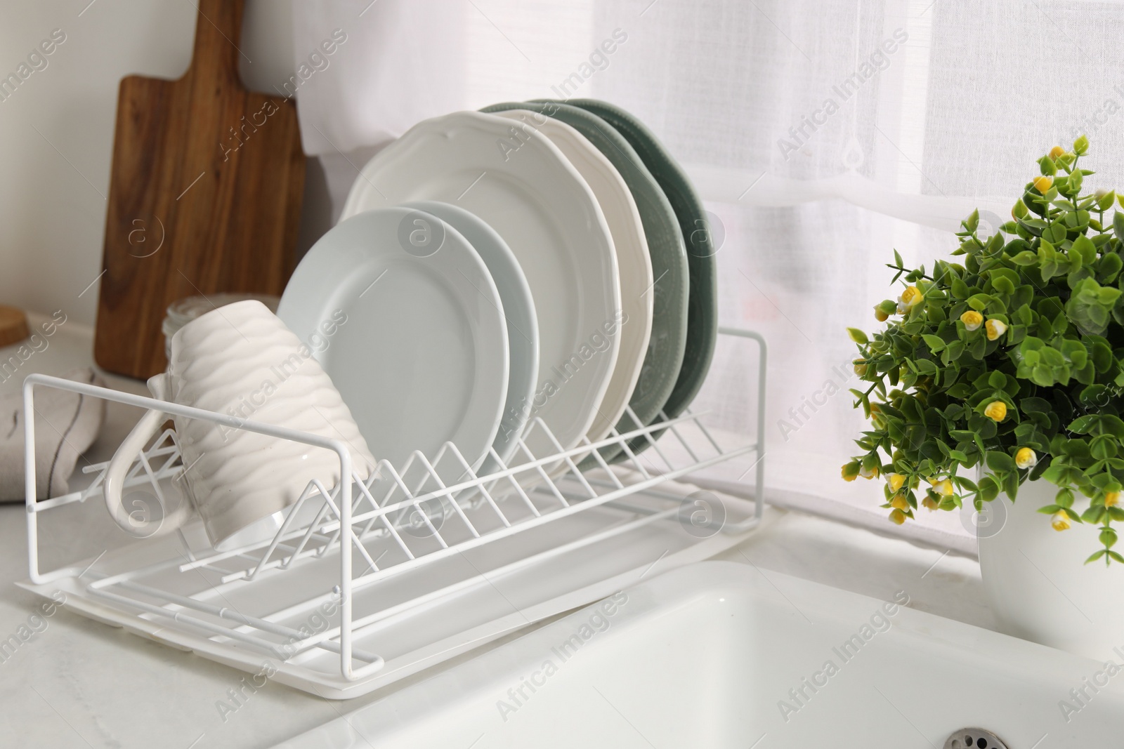 Photo of Drainer with different clean dishware, cup and houseplant on light table indoors