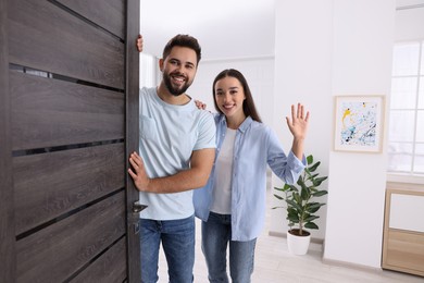 Photo of Happy couple waving near door. Invitation to come indoors