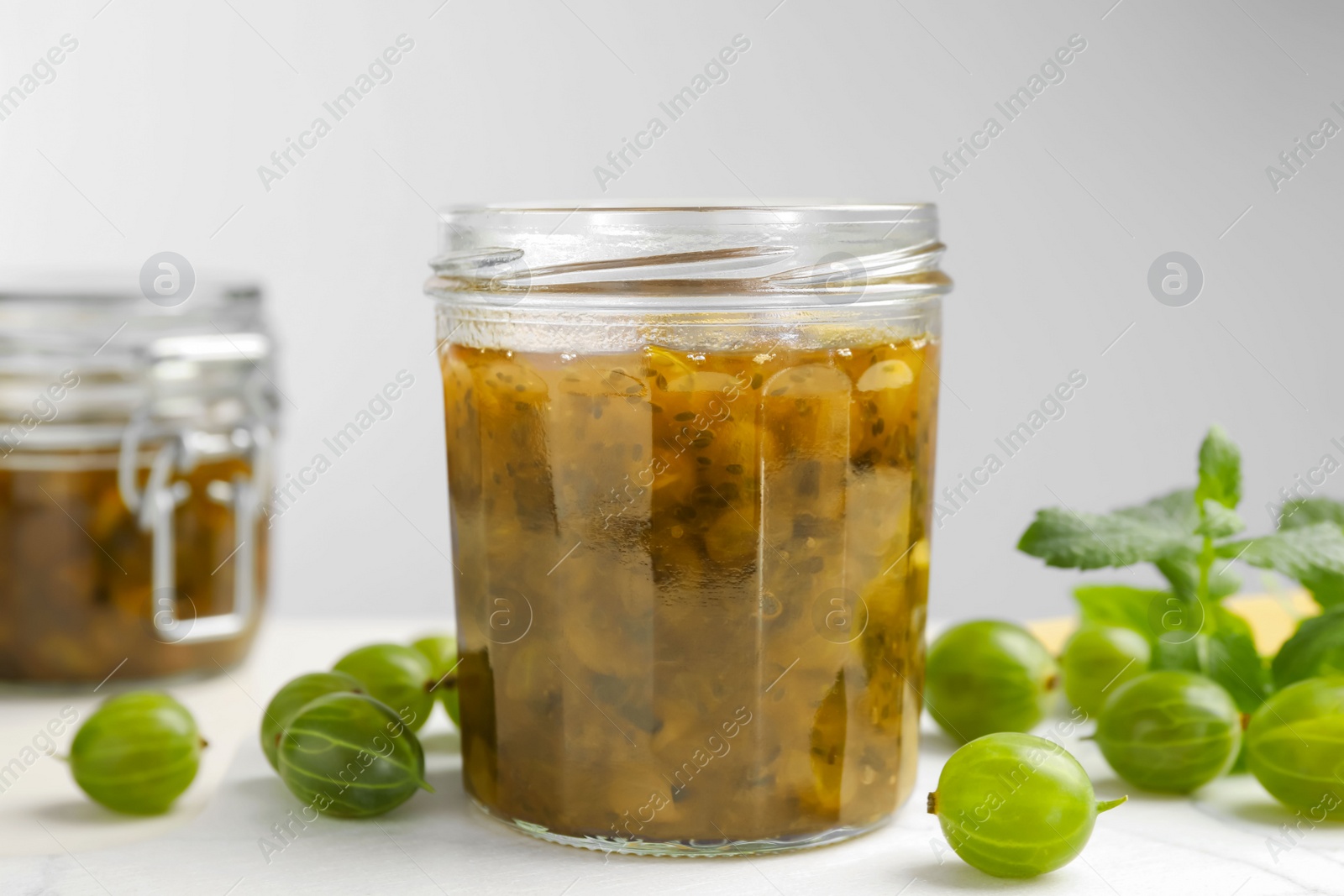 Photo of Jars of delicious gooseberry jam and fresh berries on white table, closeup