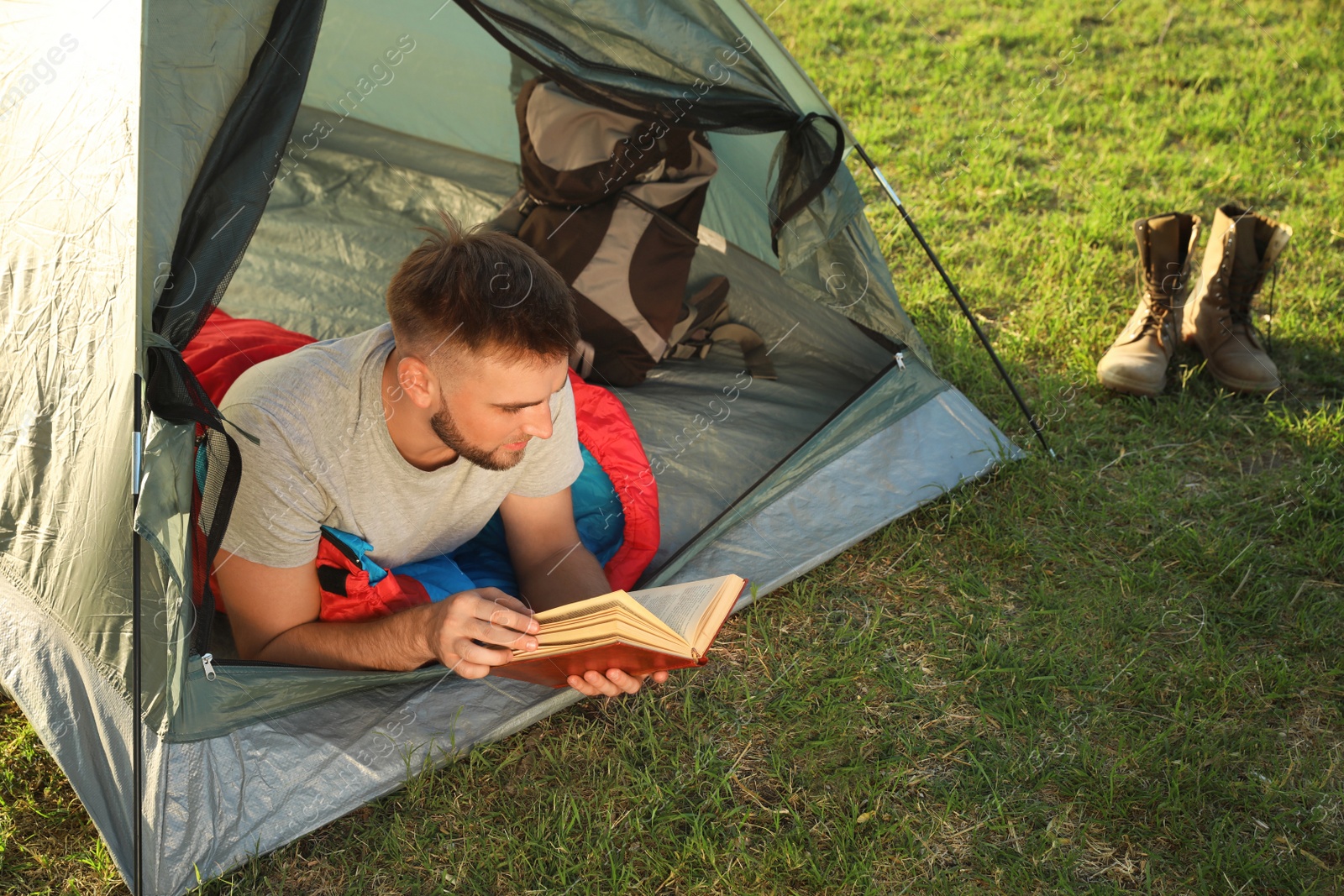 Photo of Young man in sleeping bag with book lying inside camping tent