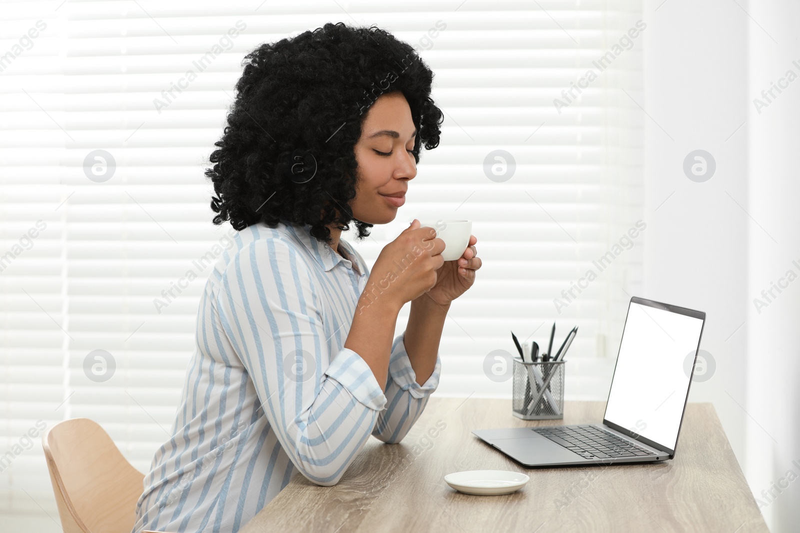 Photo of Happy young woman with cup of drink using laptop at wooden desk indoors