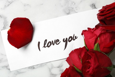 Card with text I Love You and red roses on white marble table, above view