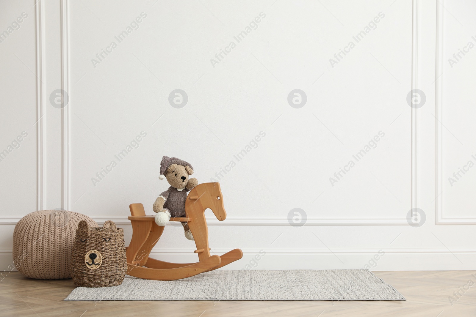 Photo of Rocking horse with bear toy, pouf and wicker basket near white wall in child room, space for text. Interior design