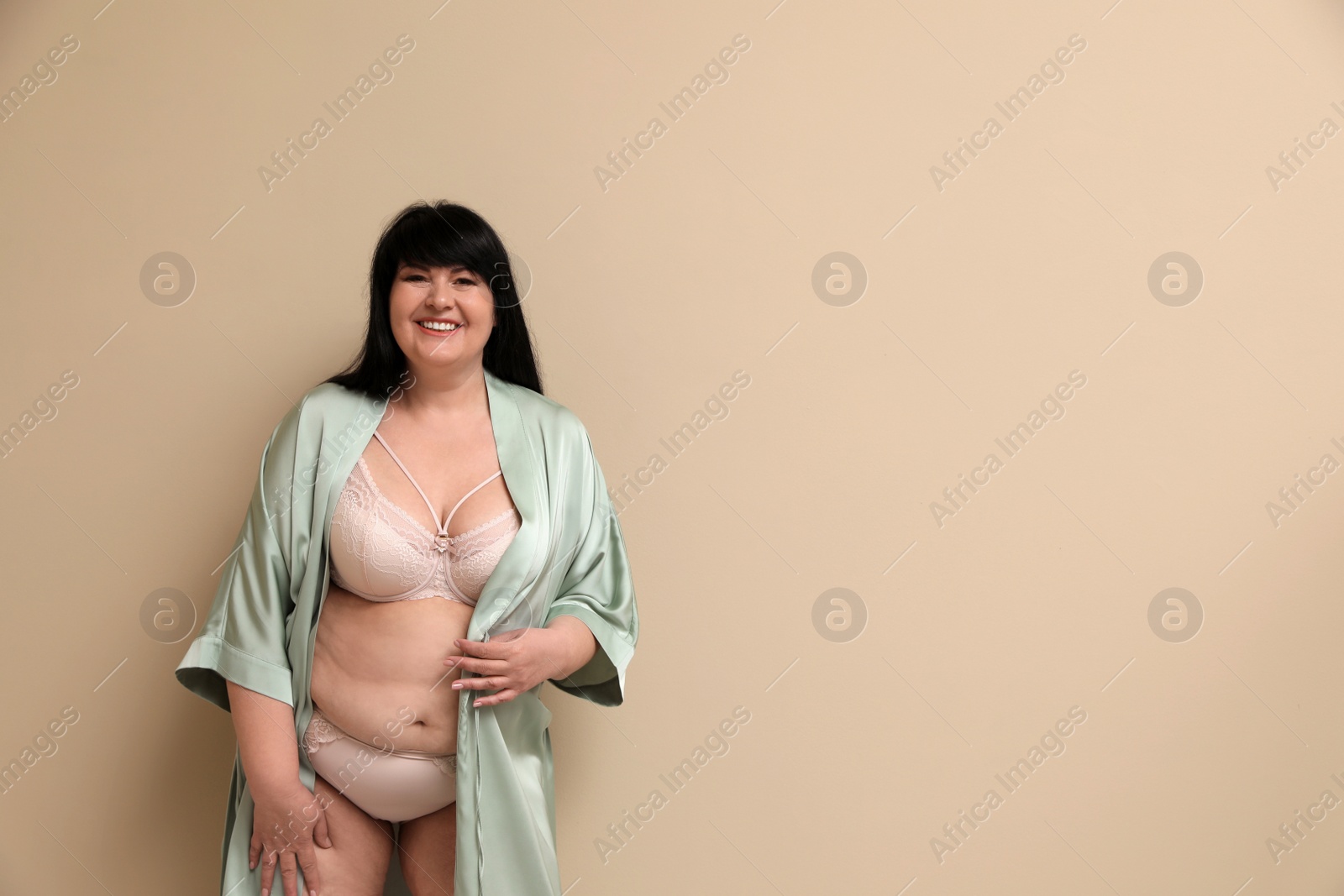 Photo of Beautiful overweight woman in sexy underwear and silk robe on beige background, space for text. Plus-size model