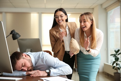 Photo of Young women popping paper bag their behind sleeping colleague in office. Funny joke