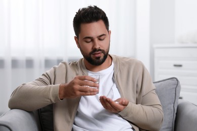 Photo of Man with glass of water taking antidepressant pill in armchair at home