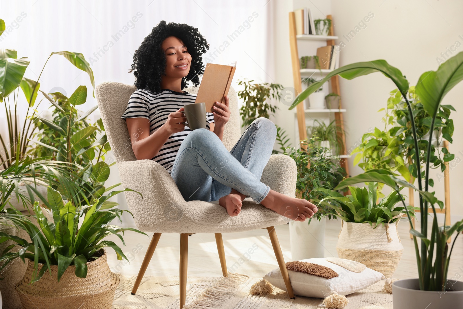 Photo of Relaxing atmosphere. Happy woman with cup of hot drink reading book in armchair surrounded by houseplants at home