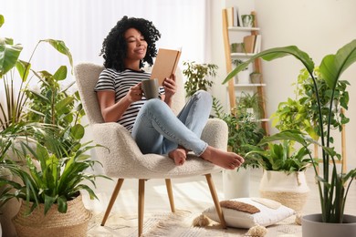 Relaxing atmosphere. Happy woman with cup of hot drink reading book in armchair surrounded by houseplants at home