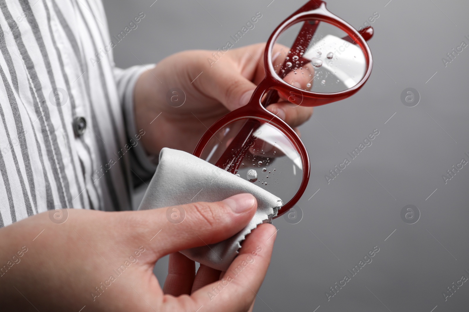 Photo of Woman wiping glasses with microfiber cloth on grey background, closeup