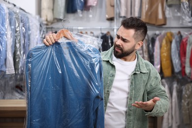 Photo of Dry-cleaning service. Confused man holding hanger with sweatshirt in plastic bag indoors