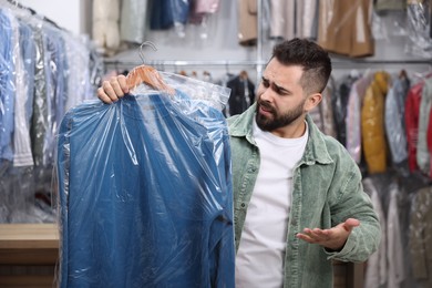 Dry-cleaning service. Confused man holding hanger with sweatshirt in plastic bag indoors