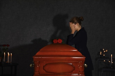 Sad young woman mourning near funeral casket with red roses in chapel