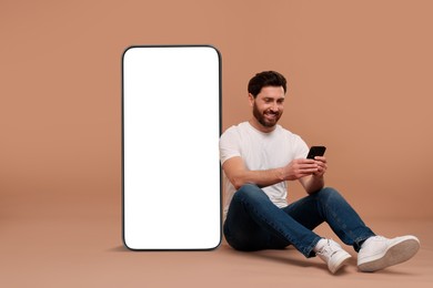 Image of Man with mobile phone sitting near huge device with empty screen on dark beige background. Mockup for design