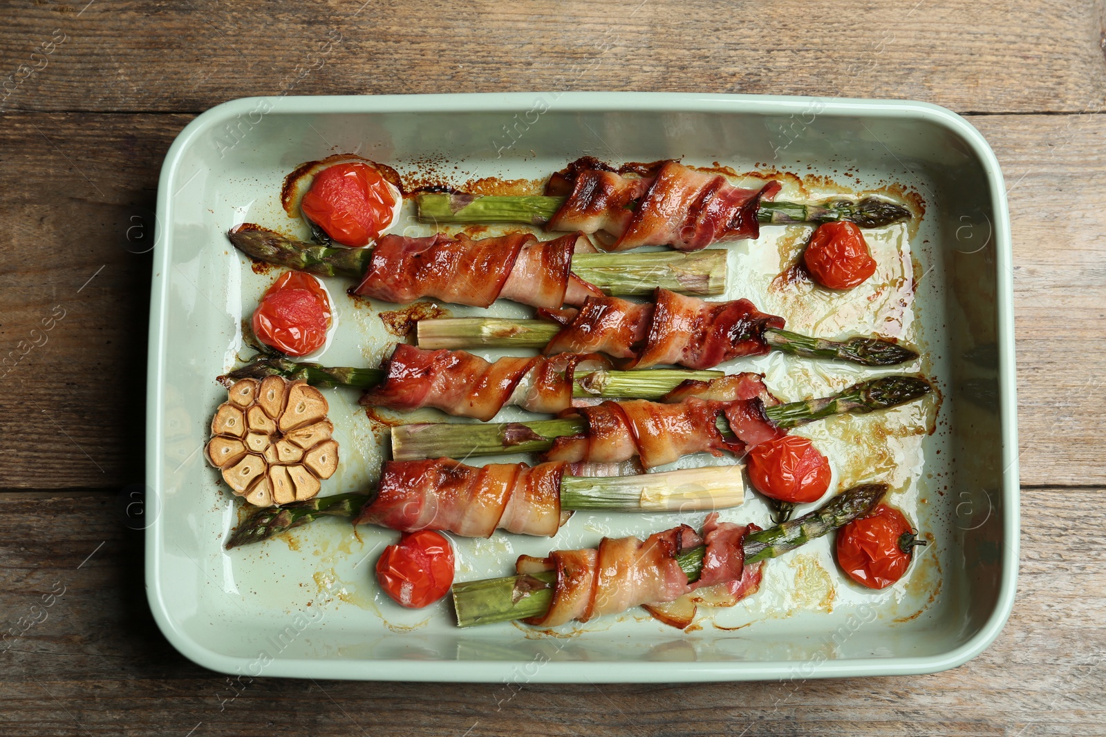 Photo of Oven baked asparagus wrapped with bacon in ceramic dish on wooden table, top view