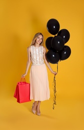 Photo of Happy young woman with balloons and shopping bags on yellow background. Black Friday Sale