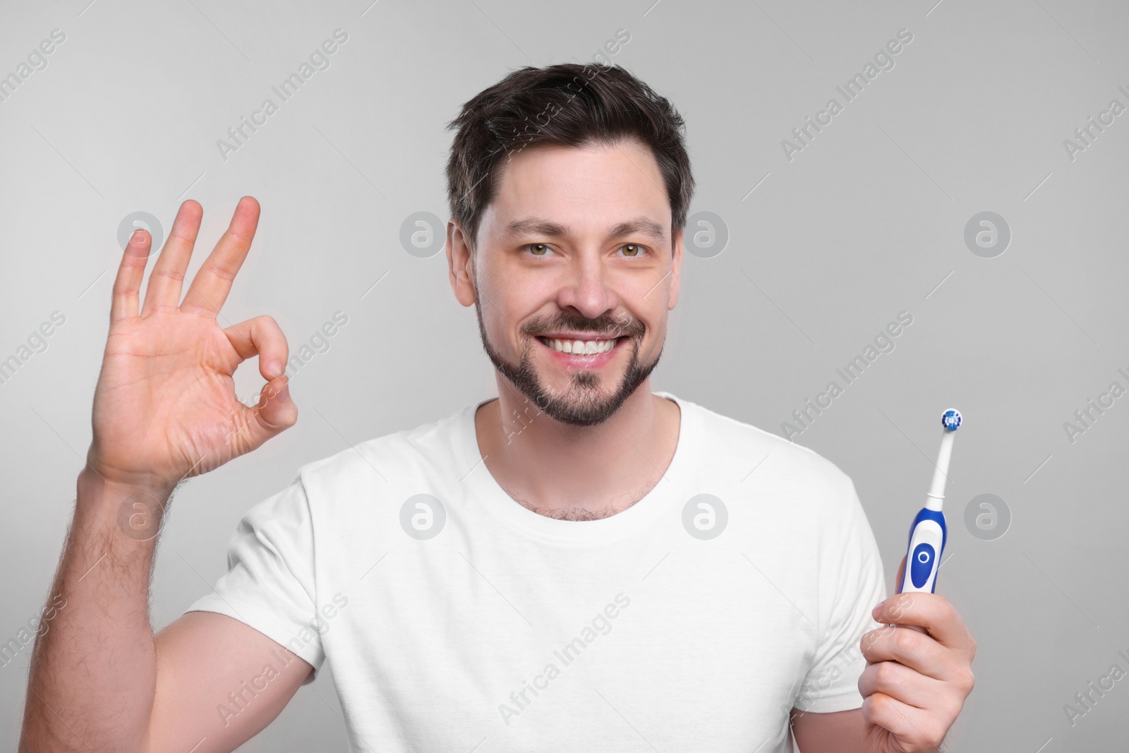 Photo of Happy man holding electric toothbrush and showing ok gesture on light grey background