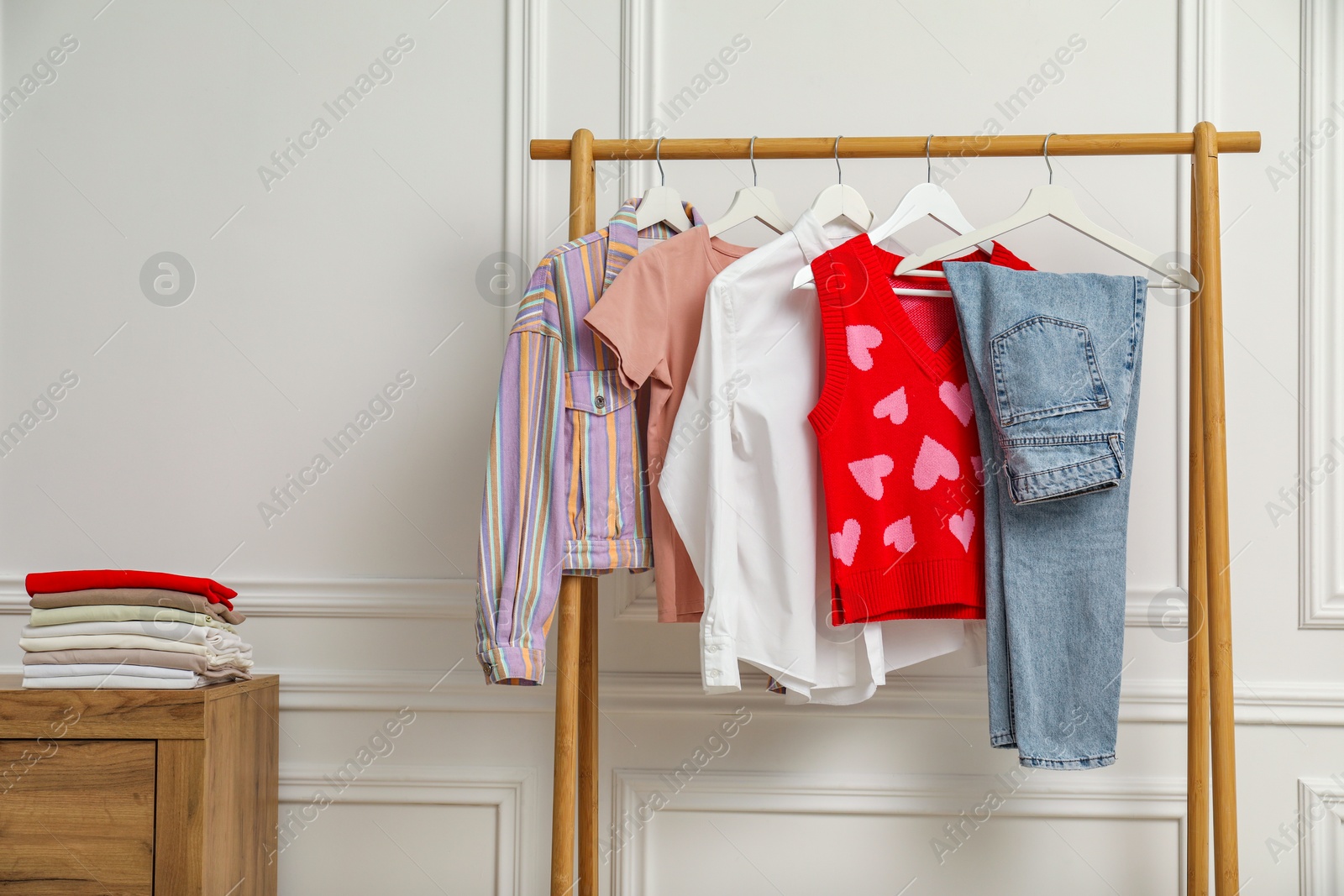 Photo of Wardrobe organization. Rack with different stylish clothes and chest of drawers near white wall