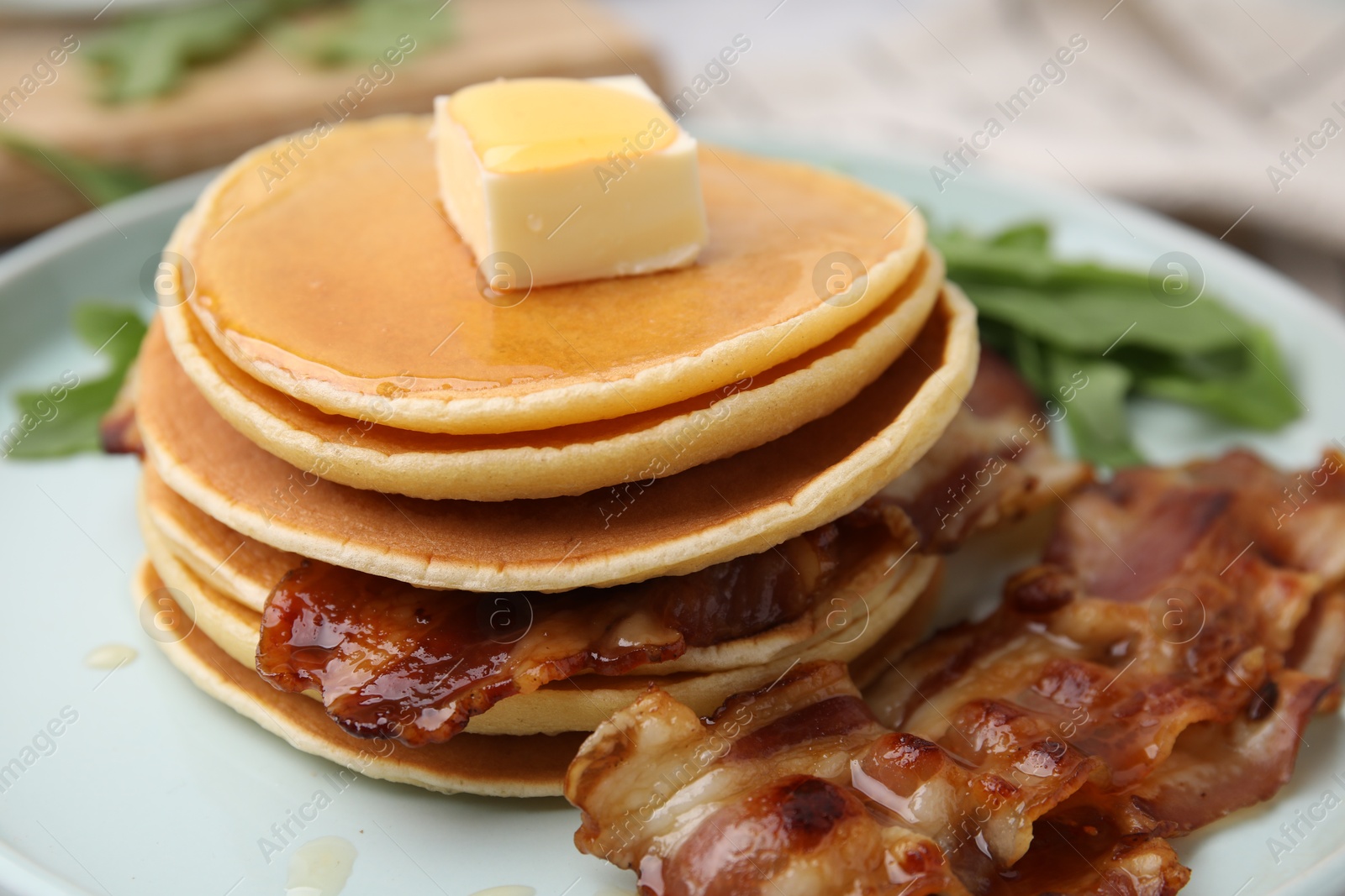 Photo of Tasty pancakes with butter, fried bacon and fresh arugula on white marble table, closeup