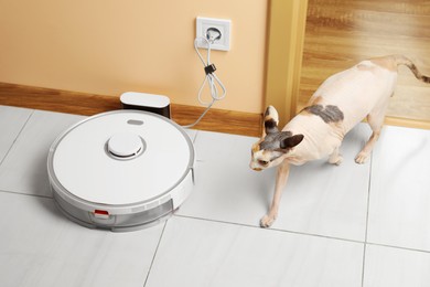 Photo of Robotic vacuum cleaner charging near beige wall and cute Sphynx cat indoors