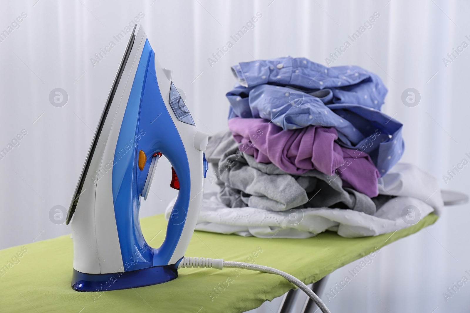 Photo of Modern electric iron and heap of clothes on board indoors. Laundry day