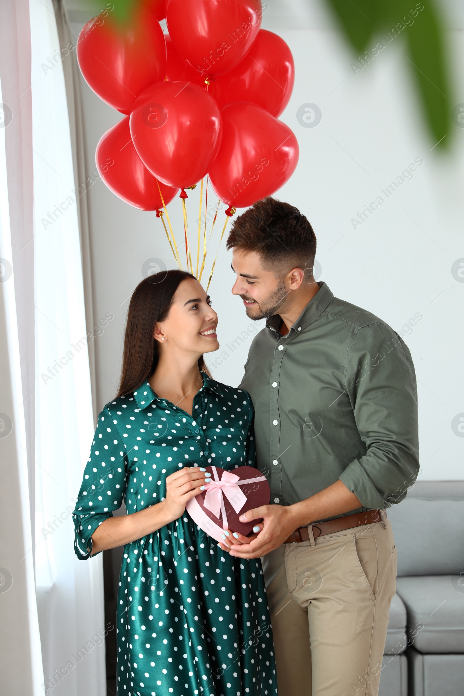 Photo of Young couple with air balloons and gift box at home. Celebration of Saint Valentine's Day