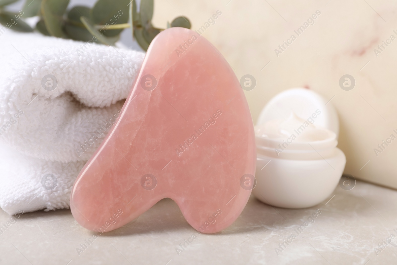 Photo of Rose quartz gua sha tool, jar of cream and towel on grey table, closeup