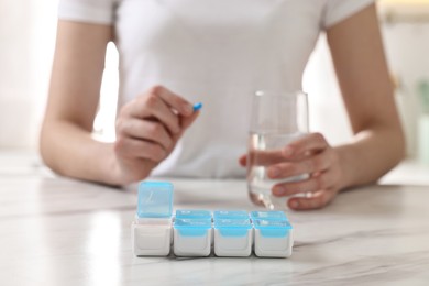 Woman with pills, organizer and glass of water at white marble table, selective focus