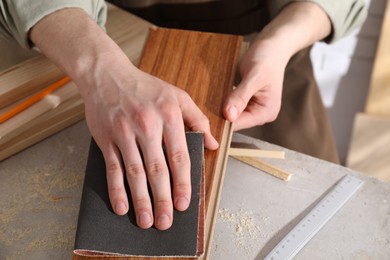 Man polishing wooden plank with sandpaper at grey table indoors, closeup
