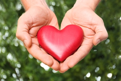 Young man holding red heart on blurred green background, closeup. Donation concept