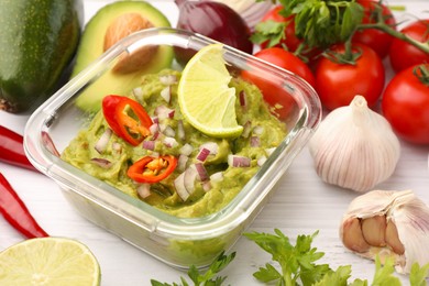 Photo of Bowl of delicious guacamole with onion, lime, chili pepper and ingredients on white wooden table, closeup