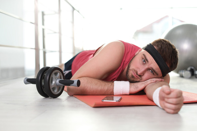 Lazy young man with abs roller lying on yoga mat indoors