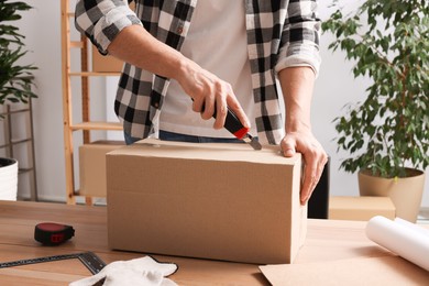 Photo of Man using utility knife to open parcel at wooden table indoors, closeup