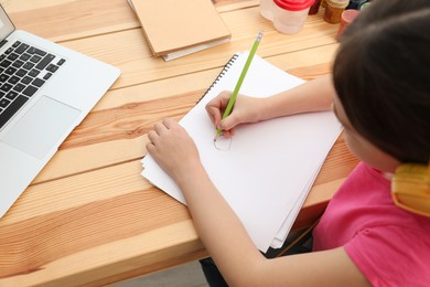 Photo of Little girl with headphones drawing on paper at online lesson indoors, closeup. Distance learning