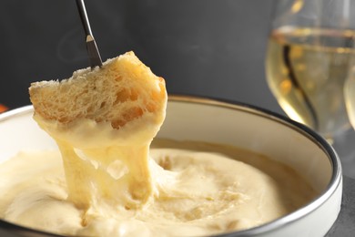 Photo of Dipping piece of bread into fondue pot with melted cheese on grey background, closeup