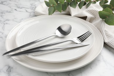Photo of Stylish setting with cutlery, napkin, eucalyptus branches and plates on white marble table, closeup