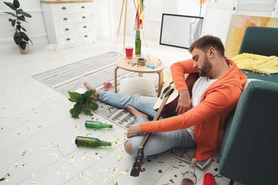 Photo of Young man with guitar sleeping near sofa in messy room after party