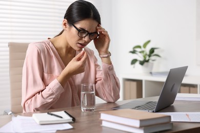 Photo of Young woman with pill and glass of water suffering from headache at wooden table in office