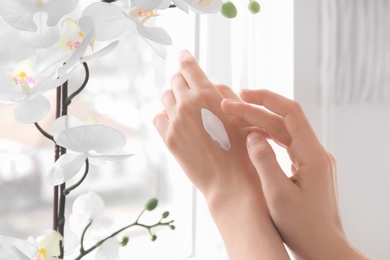 Photo of Young woman applying cream onto hands against window