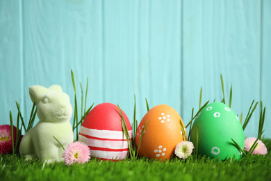 Photo of Colorful Easter eggs, rabbit and daisy flowers in green grass against light blue background. Space for text