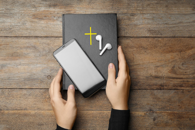 Photo of Woman with Bible, phone and earphones at wooden table, top view. Religious audiobook