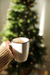 Photo of Closeup view of woman holding cocoa near Christmas  tree indoors, space for text