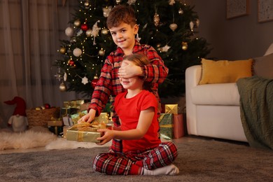 Photo of Cute little boy giving gift box to his sister near Christmas tree at home