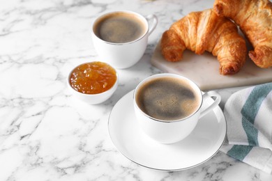 Photo of Tasty breakfast. Cups of coffee, fresh croissants and jam on white marble table, space for text