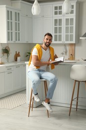 Photo of Handsome man with book sitting on stool in kitchen