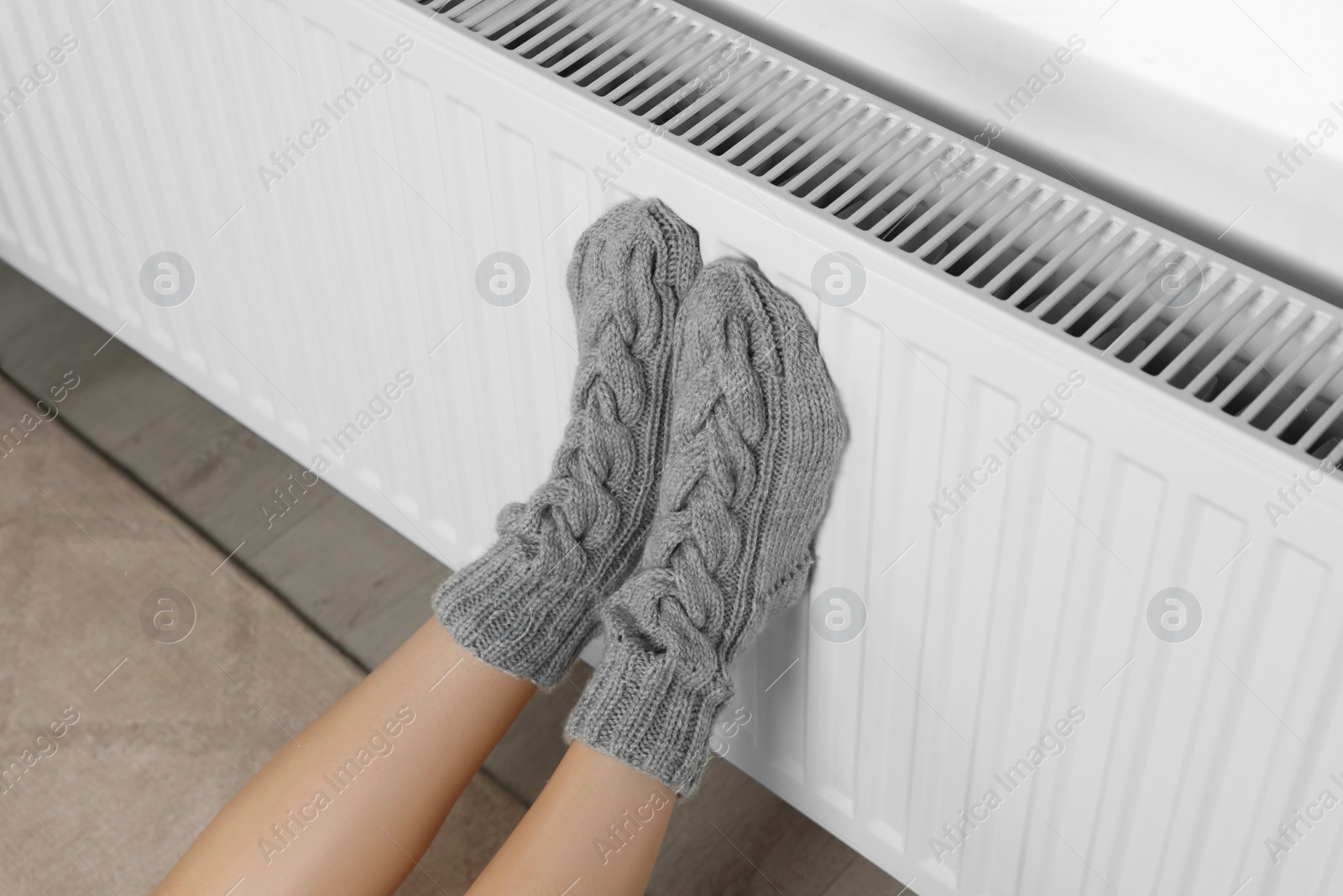 Photo of Woman warming legs on heating radiator near white wall, closeup