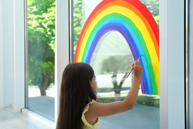 Photo of Little girl drawing rainbow on window indoors. Stay at home concept