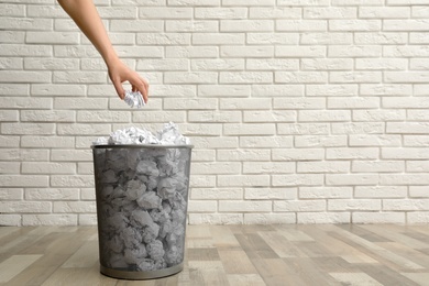 Photo of Woman throwing crumpled paper into metal bin on floor against brick wall, closeup. Space for text