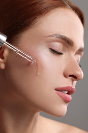 Beautiful young woman applying cosmetic serum onto her face on grey background, closeup