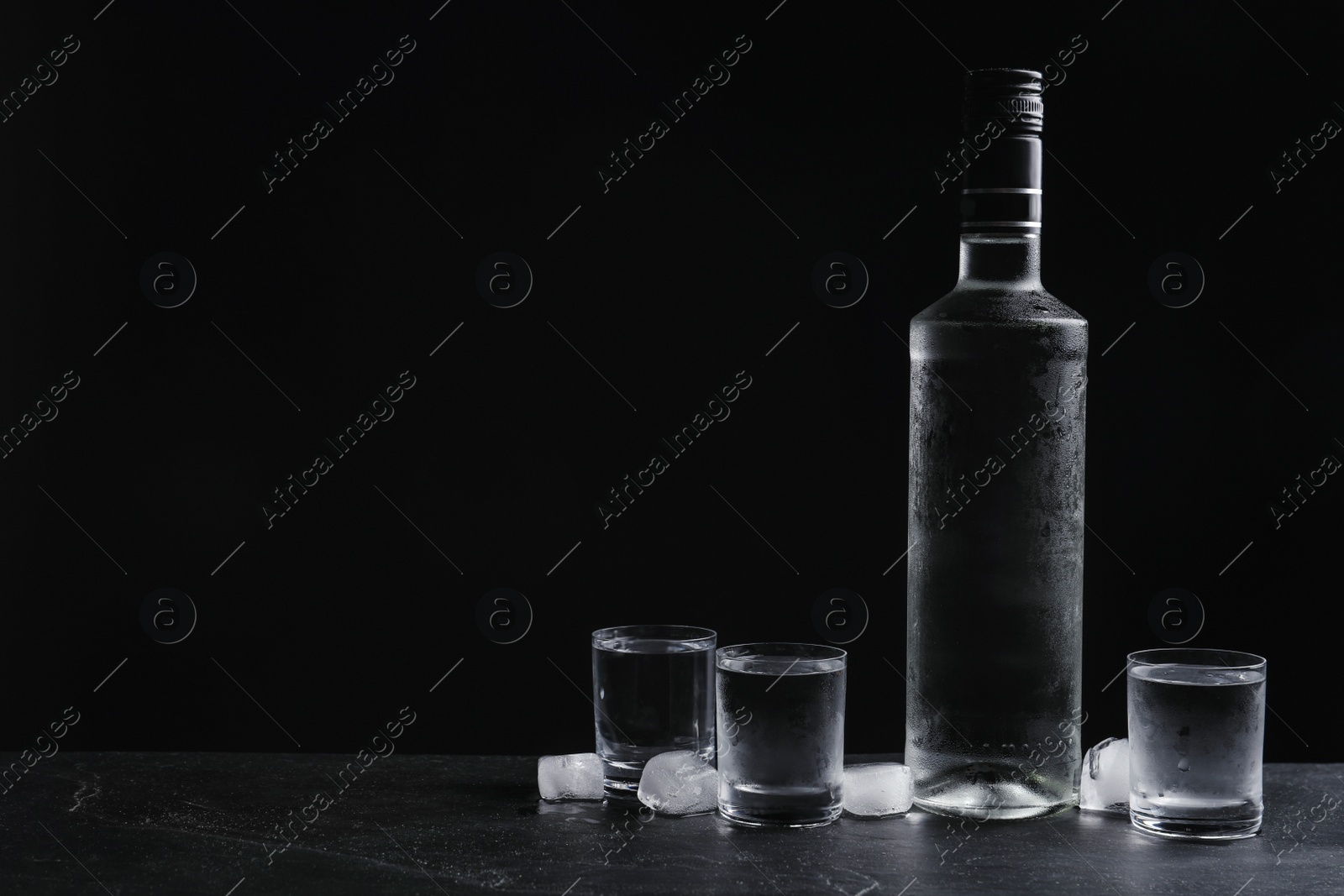 Photo of Bottle of vodka and shot glasses with ice on table against black background. Space for text